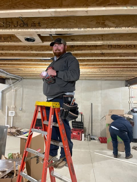 A Tom & Jerry's plumber up a step ladder with a cordless drill, running new lines through the floor joists of a basement